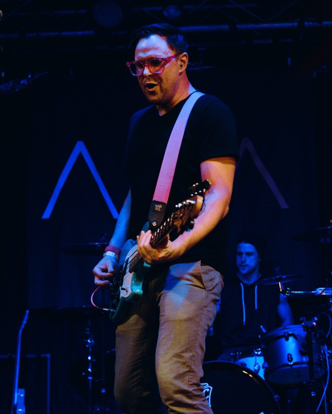 AITA guitarist intensely playing a teal electric guitar on stage, wearing glasses, with the drummer visible in the background under blue stage lights.